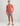 Coral short-sleeve t-shirt with small logo on chest, paired with gray shorts and brown sandals.