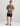 Olive green short-sleeve t-shirt with chest pocket and logo, paired with black shorts and sandals.