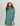 Woman in long-sleeve teal women's beach cover-up and white cap smiling in front of neutral background.