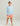 Boy wearing light blue long-sleeve crew neck shirt with HUK logo, coral shorts, and white and blue baseball cap.