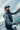 A bearded man wearing a dark gray sleeveless vest over a gray long-sleeve shirt and a navy baseball cap, holding a fishing rod on a boat with the ocean and cloudy sky in the background.