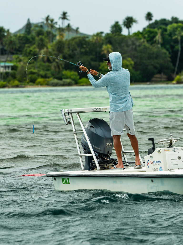 A man stands on the deck of a white fishing boat, casting a fishing rod. He is wearing a light blue long-sleeve hooded shirt and gray shorts.